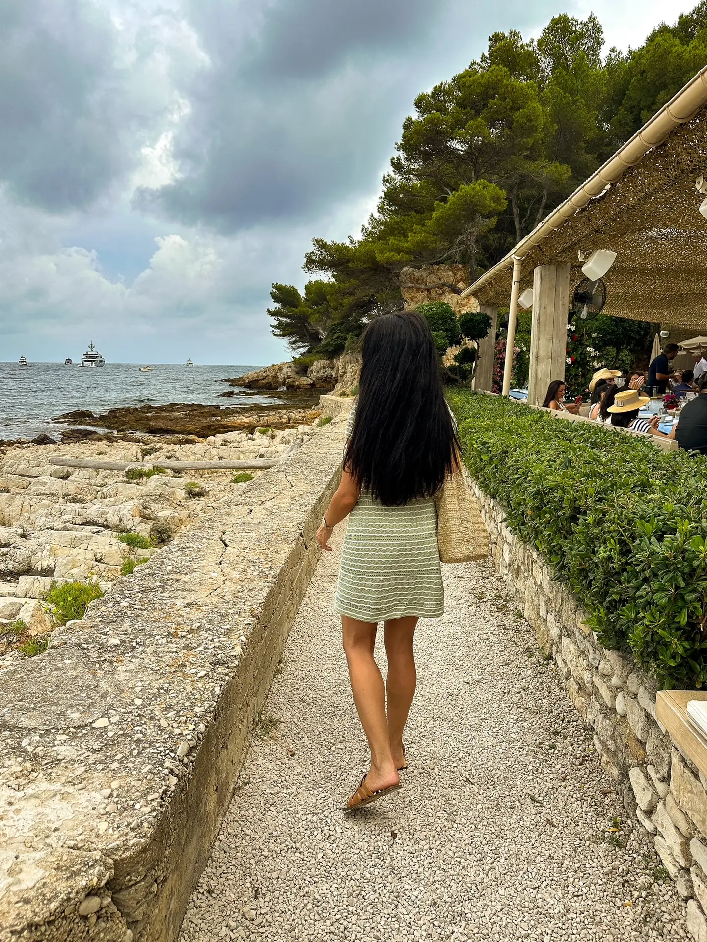 Woman walking along a sunlit path toward La Guérite Beach Club in Cannes, surrounded by Mediterranean greenery